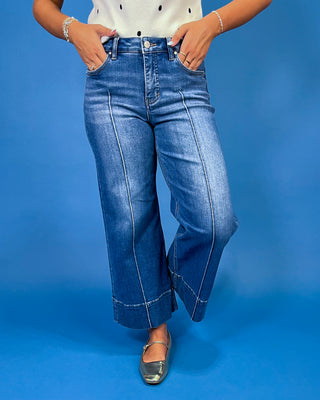 A person wearing blue wide-leg cropped jeans with rolled hems, paired with metallic silver flats, standing against a solid blue studio background.