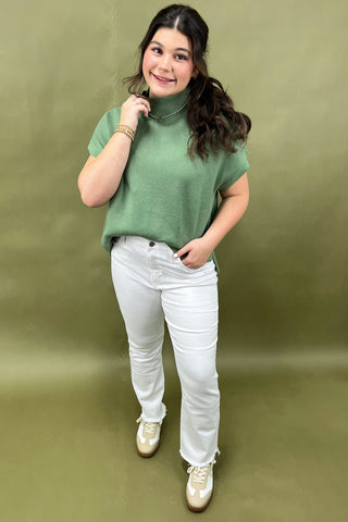 Woman wearing a green sweater and white pants against an olive green background