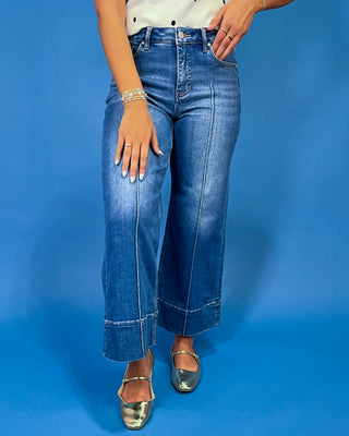 A person wearing blue wide-leg cropped jeans with a white top and metallic silver shoes, standing against a solid blue studio background.
