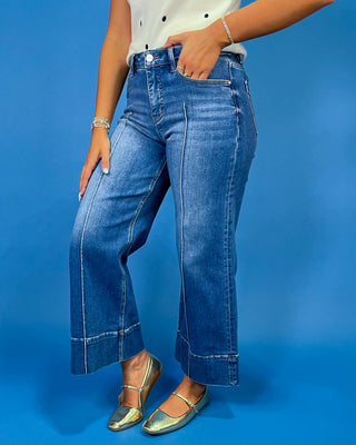 A person wearing medium-wash blue wide-leg cropped jeans with contrast stitching, paired with a white top and metallic gold flats, standing against a solid blue studio background.