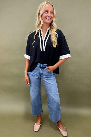 Woman wearing a black top with white trim and blue jeans against a green background