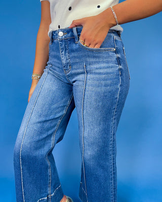 A person wearing blue denim jeans with a white top, standing against a solid blue background. The photo is cropped from the chest down, showing the front pockets, waistband, and front legs of the jeans.