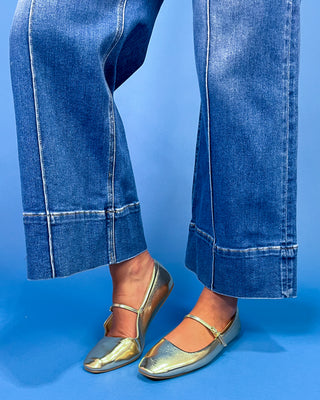 A person wearing blue wide-leg cropped jeans with rolled hems, paired with metallic silver flats, standing against a solid blue studio background.
