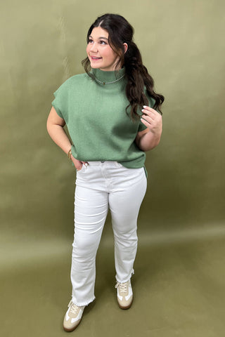 Woman wearing a green sweater and white pants against an olive green background
