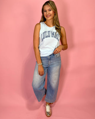 Woman wearing a white sleeveless top with 'Lulu Mac' text and blue jeans on a pink background