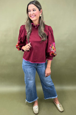 Woman wearing a burgundy blouse with floral embroidery and blue jeans on a green background
