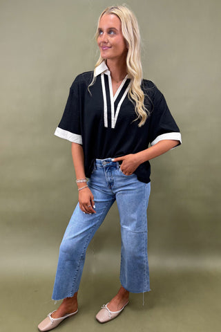 Woman wearing a black top with white accents and blue jeans against a green background