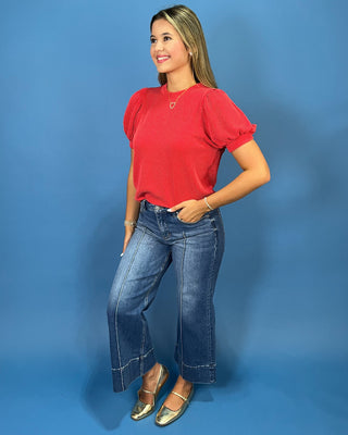 A woman modeling a red short-sleeve ribbed top with blue wide-leg jeans and metallic gold flats, standing against a solid blue studio backdrop.