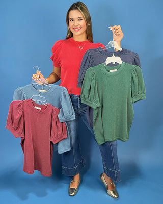 A woman stands against a blue studio background holding up four ribbed short-sleeve tops on hangers, each in a different color: red, light blue, green, and navy.