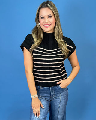 A woman modeling a black ribbed short-sleeve mock neck top with horizontal beige stripes, paired with blue jeans, against a solid blue background.