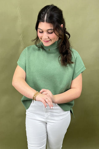 Woman wearing a green sweater and white pants against an olive green background