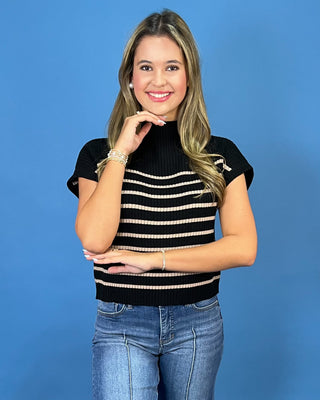 A woman modeling a black short-sleeve knit top with horizontal beige stripes, paired with blue jeans, against a solid blue studio background.