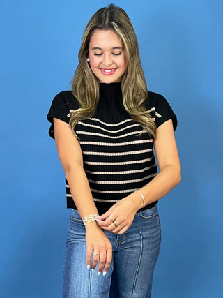 A woman with blonde-highlighted hair wearing a black short-sleeve mock neck top with horizontal beige stripes and bow details on the sleeves, paired with blue jeans, standing against a solid blue background.