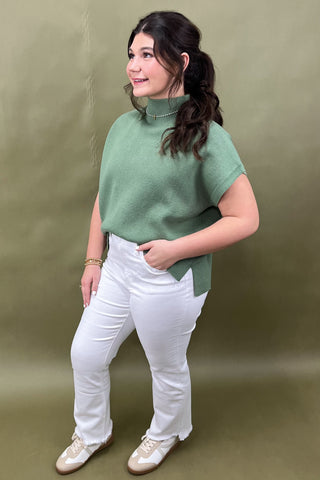 Woman wearing a green sweater and white pants against a plain background