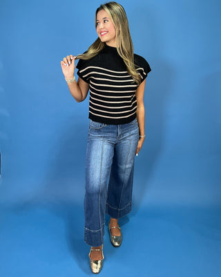 A woman modeling a black short-sleeve knit top with thin beige horizontal stripes, paired with wide-leg blue jeans and metallic gold flats, set against a solid blue studio backdrop.