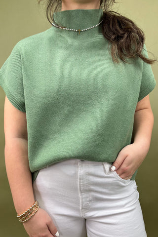 Person wearing a green knitted top and white pants against a beige background