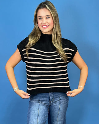 A woman modeling a black knit mock-neck top with horizontal cream stripes, paired with blue jeans, against a solid blue background.