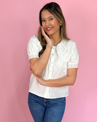Woman wearing a white blouse with lace details and blue jeans against a pink background