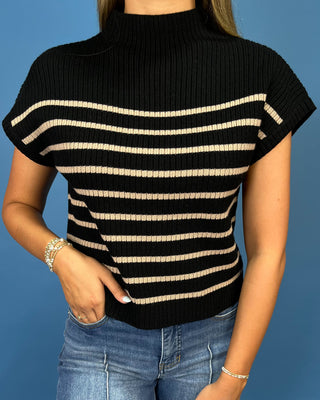 A woman wearing a black ribbed short-sleeve mock neck top with horizontal beige stripes, paired with blue jeans, against a solid blue background.
