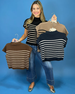 A woman stands against a solid blue studio background holding three ribbed knit mock-neck crop tops on hangers. The tops are shown in brown and beige with white horizontal stripes, and she is also wearing a black mock-neck top with white stripes and blue jeans.