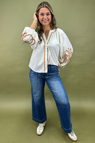 Woman wearing a white blouse with brown embroidery and blue jeans on a green background