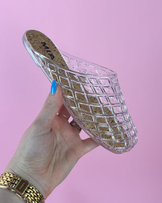 Clear glass slipper held by a hand with blue nail polish against a pink background