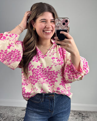 Woman taking a mirror selfie wearing a pink floral blouse and blue jeans.