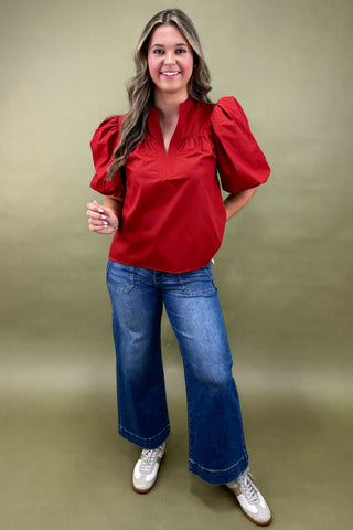 Woman wearing a red blouse with puffed sleeves and blue jeans on a green background
