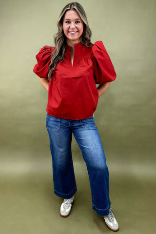 Woman wearing a red blouse with puff sleeves and blue jeans on a green background