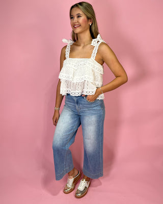 Woman wearing a white lace top and blue jeans against a pink background
