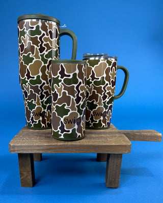 Three insulated travel tumblers with a green, brown, and beige camouflage pattern are displayed on a rustic wooden stand against a solid blue background.