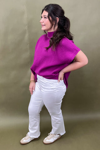Woman wearing a bright purple sweater and white pants against a plain background