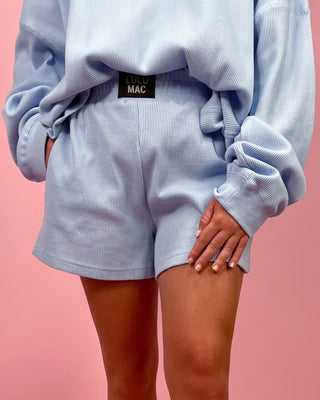 Person wearing a light blue oversized sweatshirt and shorts with a visible MAC logo on a pink background