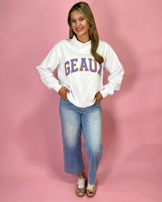 Woman wearing a white sweatshirt with 'GEAUX' text and blue jeans on a pink background