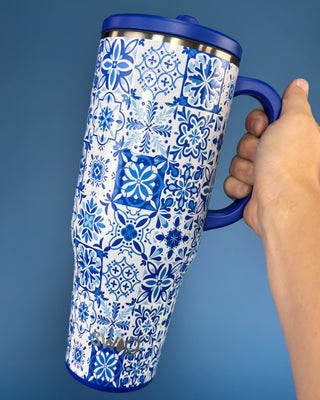 A hand holding a tall insulated travel tumbler with a blue lid and handle, featuring a white body covered in a blue floral and tile-pattern print. The brand name 'KAVU' is printed near the bottom.