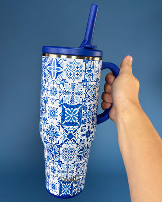 A hand holding a tall insulated tumbler with a blue lid and straw. The tumbler has a white body covered in a blue geometric tile pattern, with a blue handle and blue rim, set against a solid blue background.
