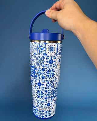 A hand holding a tall insulated tumbler with a blue handle and lid