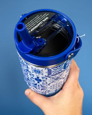 A hand holding a blue and white patterned insulated tumbler with a blue lid and straw, shown against a solid blue background.