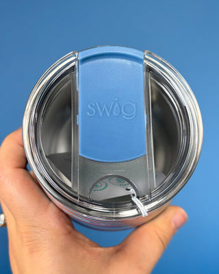 A close-up of a hand holding a stainless steel tumbler with a blue and clear Swig flip-top lid against a solid blue background.