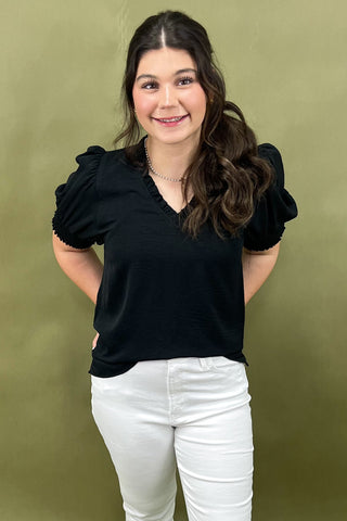 Woman wearing a black top with puff sleeves and white pants against a green background