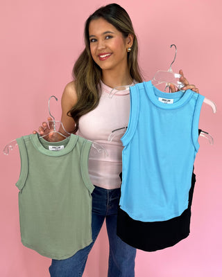 Woman holding three sleeveless tops on hangers against a pink background