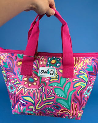 A hand holds a Swig brand tote bag with a bright pink and multicolor psychedelic pattern against a solid blue background.