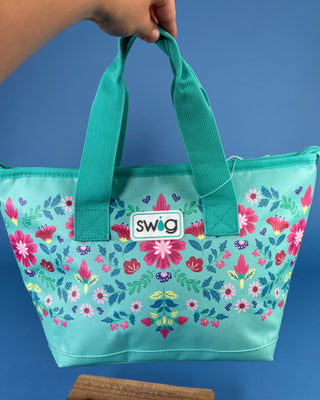 A Swig brand insulated tote bag in turquoise with a colorful floral pattern, held by a hand against a solid blue background.