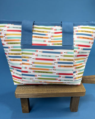 A tote bag with a colorful pencil print and motivational text, featuring blue fabric handles and trim, displayed on a small wooden stool against a solid blue background.