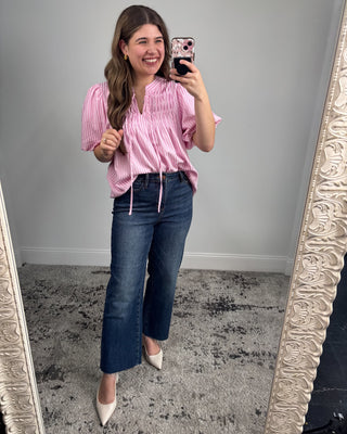 Woman taking a mirror selfie wearing a pink blouse and blue jeans.