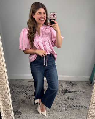 Woman taking a mirror selfie wearing a pink blouse and blue jeans.