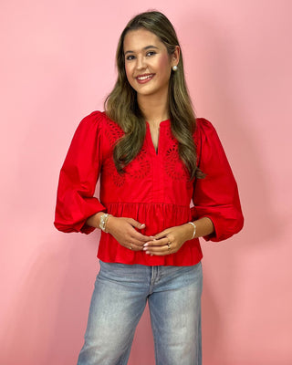 Woman wearing a red blouse with embroidery and light blue jeans against a pink background