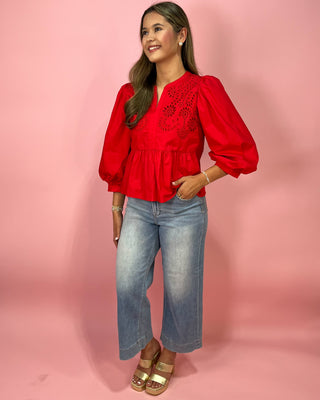 Woman wearing a red blouse with lace details and blue jeans on a pink background