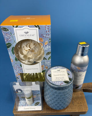 Greenleaf Home fragrance set in the scent 'Laurel Grove', including a tall floral-patterned box, a blue textured jar candle, a silver spray bottle, and a small oil bottle in a clear package, arranged on a wooden block against a blue background.