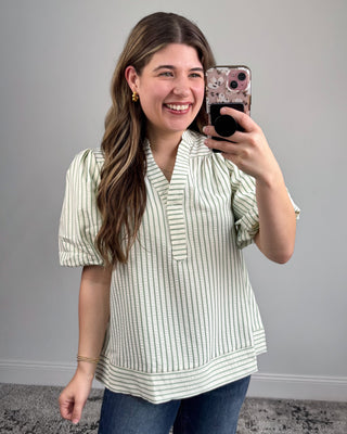 Woman taking a mirror selfie wearing a striped blouse and jeans against a gray wall.